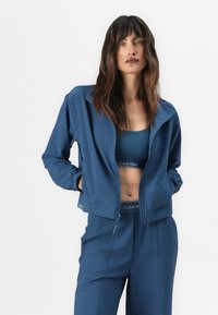Modrá sportovní bunda vyrobená z hladkého materiálu, s zapínáním na zip, elastickými manžetami a moderním střihem. Nosí se přes odpovídající sportovní podprsenku.