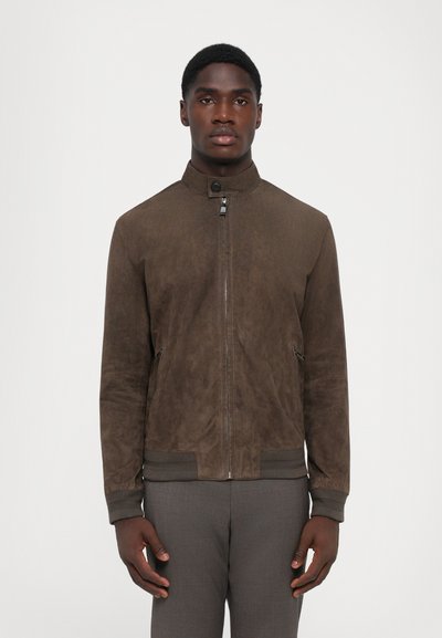 Bruine suède jas met hoge kraag, ritssluiting, zijzakken en ribgebreide manchetten. Model draagt een grijze broek. Eenvoudig en strak design.