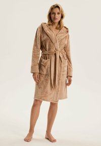 Szlafrok z kapturem uszyty z miękkiego, jasnobrązowego materiału. Posiada pasek, dwie przednie kieszenie i teksturowane mankiety. O długości średniej. Model prezentuje się boso.