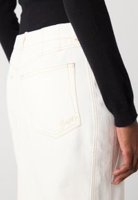 Witte denim rok met een hoge taille, voorzien van een achterzak en tan steekwerk. De zak heeft een lichte geborduurde logo.