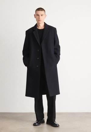 Black wool overcoat with a notched collar, button front, and side pockets; paired with black trousers and shoes. Smooth texture, tailored fit.