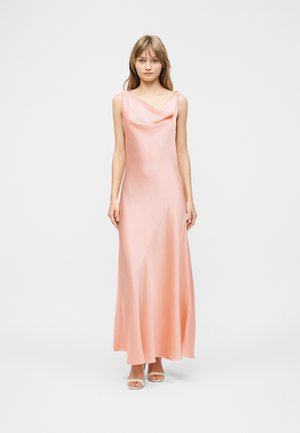 Ung kvinde står iført en gulvlængde, ærmeløs, lyserød satin kjole med draperet halsudskæring og hvide åbne stiletter.