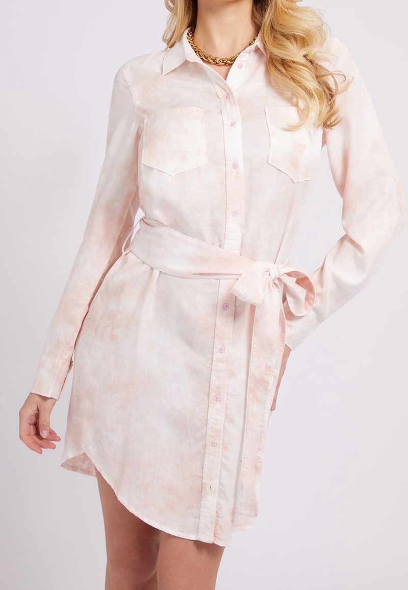 Femme portant une robe-chemise à manches longues rose pâle avec des poches poitrine et une ceinture nouée à la taille, accessoirisée d'un collier en chaîne dorée.