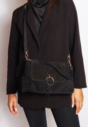 Schwarze Wildleder-Crossbody-Tasche mit strukturiertem Finish, ausgestattet mit einem goldenen Ring-Element und verstellbarem Tragegurt. Rechteckige Form mit Klappenverschluss.