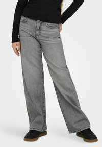 Plati pilkai džinsiniai platėjantys kelnės su išblukusiu efektu, išsamiais siūlais, vidutinio aukščio liemeniu ir priekinėmis kišenėmis, derinami su juodais sportbačiais.