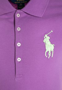 Lila polo skjorta i texturerad tyg med tre knappar och en broderad ljusgrön polo-spelare logotyp på vänster bröst.