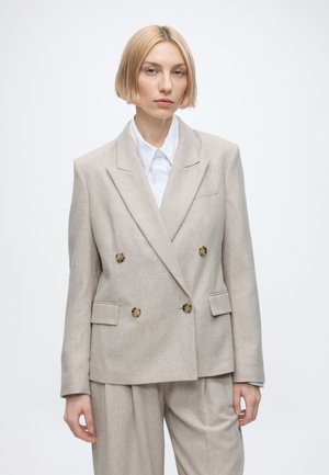 Ung kvinde med kort blondt hår bærer en beige dobbeltradet blazer og matchende bukser over en hvid skjorte mod en ensfarvet baggrund.