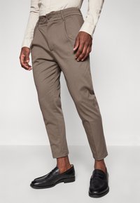 Pantalones marrones ajustados con pierna entallada, que cuentan con un cierre frontal de botón y detalles de pliegues. Combinados con mocasines negros.