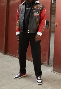 Czarna skórzana kurtka bomberka z czerwonymi rękawami i wieloma naszywkami, w połączeniu z czarną bluzą z kapturem i czarnymi dżinsami, uzupełniona czarno-czerwonymi sneakersami.