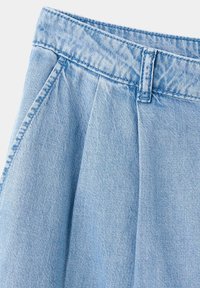 Jupe en denim bleu clair avec passants de ceinture, plis et poches latérales, visible de la taille jusqu'à mi-cuisse.