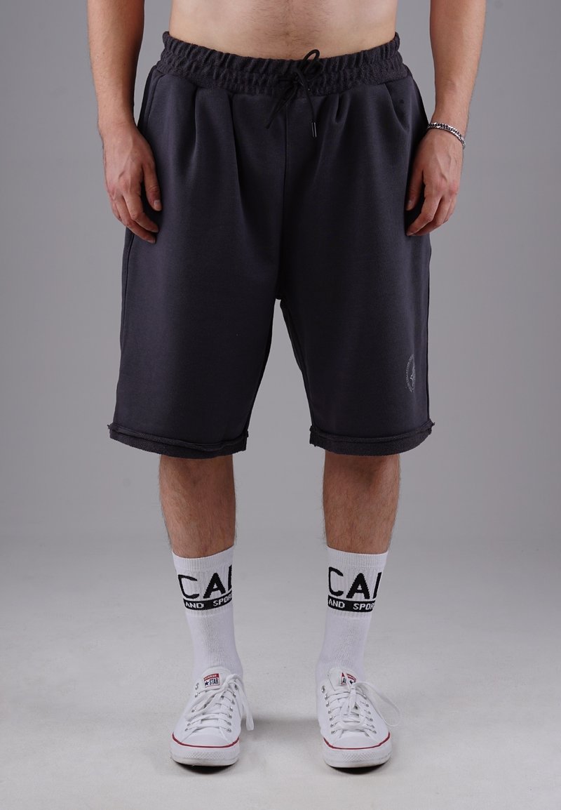 Dunkelgraue Sweatshorts mit elastischem Bund und Kordelzug. Sie bieten eine lockere Passform mit umgeschlagenen Säumen. Kombiniert mit weißen Socken und Sneakern.