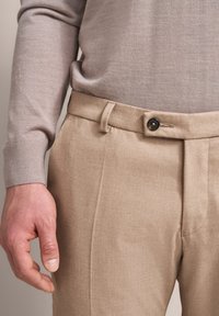 Beige Taillierte Hose zeigt eine glatte Textur, eine flache Vorderseite und einen einzelnen Knopfverschluss. Die Hand des Models liegt nahe dem Bund.