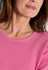 Nahaufnahme einer Frau, die einen strukturierten rosafarbenen Pullover und glänzenden rosafarbenen Lippenstift trägt, wobei ihr Hals und der untere Teil ihres Gesichts zu sehen sind.