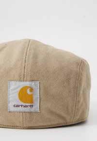 Gorra de tela color beige con una superficie texturizada, que presenta un parche de logo cuadrado en gris y naranja con "Carhartt" impreso en rojo oscuro.