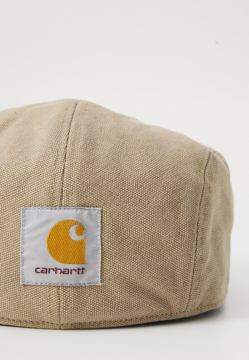 Beige stoffen pet met een gestructureerd oppervlak, voorzien van een vierkant logo-patch in grijs en oranje met daarop "Carhartt" in donkerrood gedrukt.