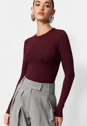 Bordowa bluzka z długim rękawem wykonana z elastycznego materiału, z okrągłym dekoltem i dopasowanym krojem, zestawiona z wysokim stanem szarymi spodniami.