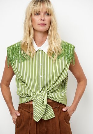 Groen gestreept overhemd met franjes op de schouders, met kraag en knoopsluiting. Geknoopt in de taille, gecombineerd met een bruine broek met hoge taille.