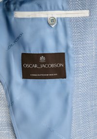 Tessuto azzurro chiaro con una texture a spina di pesce; presenta un'etichetta marrone con "Oscar Jacobson" e dettagli sulla lavorazione svedese.