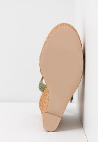 Sandale avec semelle et talon tissés couleur beige, bride tressée verte, vue du côté de la semelle sur fond blanc.