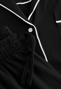 Completo pigiama nero composto da una maglietta con bottoni, rifiniture bianche e tasca, e pantaloni con vita elastica e dettaglio con cordino. Tessuto morbido.