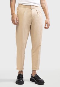 Pantalons en coton beige avec une finition texturée, dotés de plis sur le devant et de bas roulés. Portés avec des mocassins noirs.