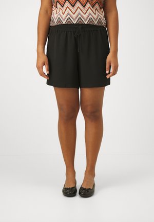 Frau trägt schwarze Shorts mit elastischem Bund, gemustertes ärmelloses Oberteil und schwarze Ballerinas, steht vor einem einfarbigen Hintergrund.