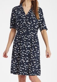 Kvinna i en marinblå klänning med korta ärmar och ett litet blommönster i vitt, beige och ljusblått, stående med avslappnade armar.