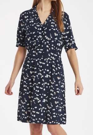 Femme portant une robe bleu marine à manches courtes avec un petit motif floral en blanc, beige et bleu clair, debout, les bras relâchés.