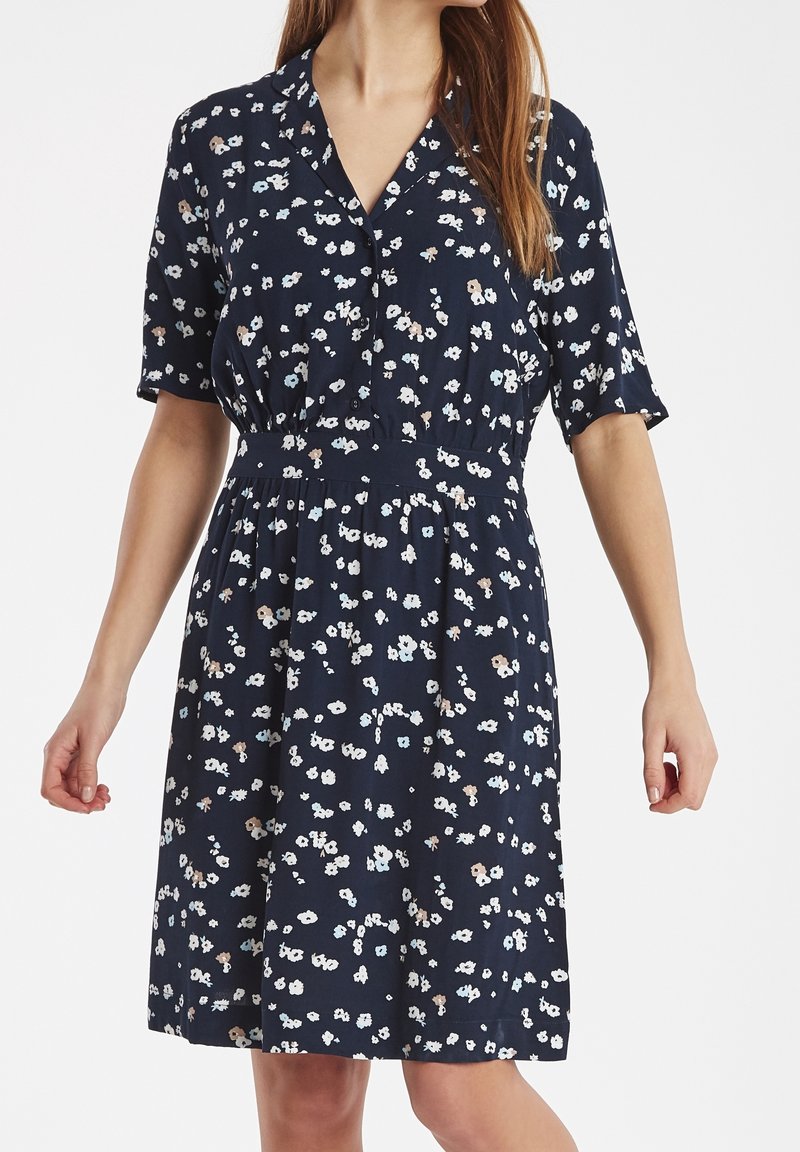 Kvinna i en marinblå klänning med korta ärmar och ett litet blommönster i vitt, beige och ljusblått, stående med avslappnade armar.