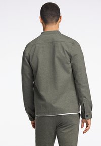 Chaqueta texturizada de color verde oscuro con un cuello estructurado, puños abotonados y corte recto; llevada sobre pantalones a juego, vista desde la parte de atrás.