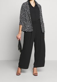 Combinaison noire à jambes larges associée à un blazer noir et blanc à motifs. Le blazer présente un imprimé léopard, et le mannequin tient une pochette noire.