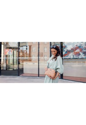 Tan leather shoulder bag with circular cut-out pattern and zipper closure. The bag is held by a person in a light green dress, outdoors.