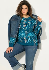 Felpa verde acqua con motivo floreale, dal collo rotondo e polsini a coste, abbinata a jeans blu e a una giacca di jeans. Scarpe color argento.
