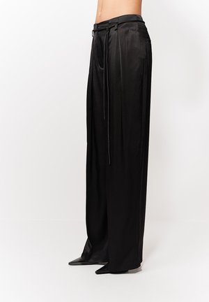 Juodos plačiomis kelnėmis iš satino su diržo kilpomis ir ilga raišteliu, dėvimos su juodais aštriais bateliais, matomos nuo juosmens žemyn.