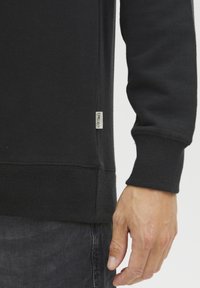 Black sweatshirt featuring a ribbed hem and cuffs, with a small label on the side seam. The fabric appears soft and thick, indicating warmth.