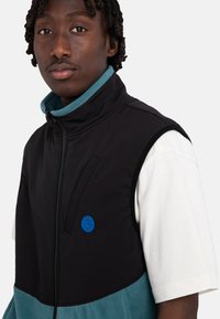 Black and teal sleeveless vest with a high collar, front zipper, and prominent blue logo detail. Soft, textured fabric. White shirt underneath.