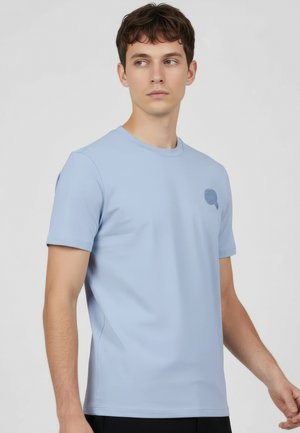 Giovane uomo che indossa una maglietta a maniche corte azzurra con un piccolo emblema sul petto, in piedi contro uno sfondo grigio uniforme.