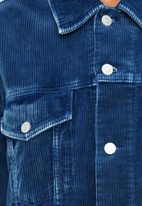 Giacca in corduroy di colore blu scuro con righe verticali, dotata di colletto a punta e bottoni a pressione argentati, e due tasche sul petto.