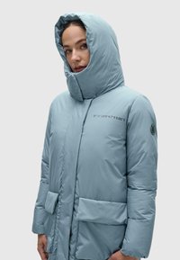 Svijetloplavi puffer kaput s visokim ovratnikom, oversized kapuljačom i prednjim džepovima. Sadrži tekstualni dizajn na prsima i logo na rukavu.