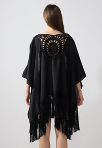 Caftán negro con un diseño circular de ganchillo en el cuello y bordes con flecos. Tela ligera que se drapea de manera suelta.