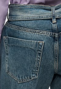 Οπίσθια όψη μεσαίου μπλε τζιν τζιν με ορατή πίσω τσέπη και θηλιά για ζώνη, φορεμένα με πουκάμισο λαβαντί που έχει μπει μέσα.