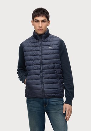 Jeune homme portant une veste matelassée sans manches bleu marine avec une fermeture éclair sur le devant et une chemise sombre à manches longues, debout devant un fond uni.