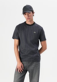 Tricou grafic negru cu un model geometric și un logo mic verde. Asortat cu blugi gri, contribuind la un aspect casual.