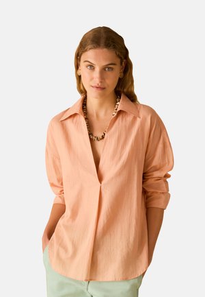 Donna con capelli castano chiaro mossi indossa una camicia oversize color pesca, collana con conchiglia, orecchini a bottone d’oro e pantaloni verde chiaro, mani nelle tasche.