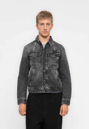 Giovane uomo che indossa una giacca di denim nera sbiadita e pantaloni neri, in piedi contro uno sfondo bianco semplice, rivolto in avanti.