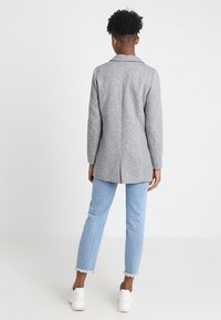 Kvinna med kort lockigt hår som bär en grå lång kavaj, ljusblå avklippta jeans med fransade kanter och vita sneakers, stående mot vit bakgrund.