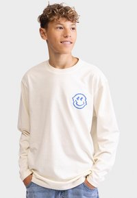 Cream long-sleeve t-shirt featuring a blue smiley face graphic on the left chest, made from soft cotton fabric, with a relaxed fit.