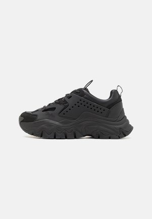 Black sneakers made of smooth and textured materials. Features thick, wavy soles, perforated panels, and lace-up design. Accents include a small pull tab.