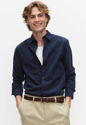 Ung mand smilende, iført navyblå skjorte over hvid t-shirt, beige bukser med flettet brunt bælte, hænder i lommerne, mod hvid baggrund.