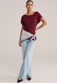 Teksturirana bordova majica s kratkimi rokavi, beli šal z pikami zavezan okoli pasu, kombiniran z svetlo modrimi širokimi kavbojkami in črnimi koničastimi čevlji.